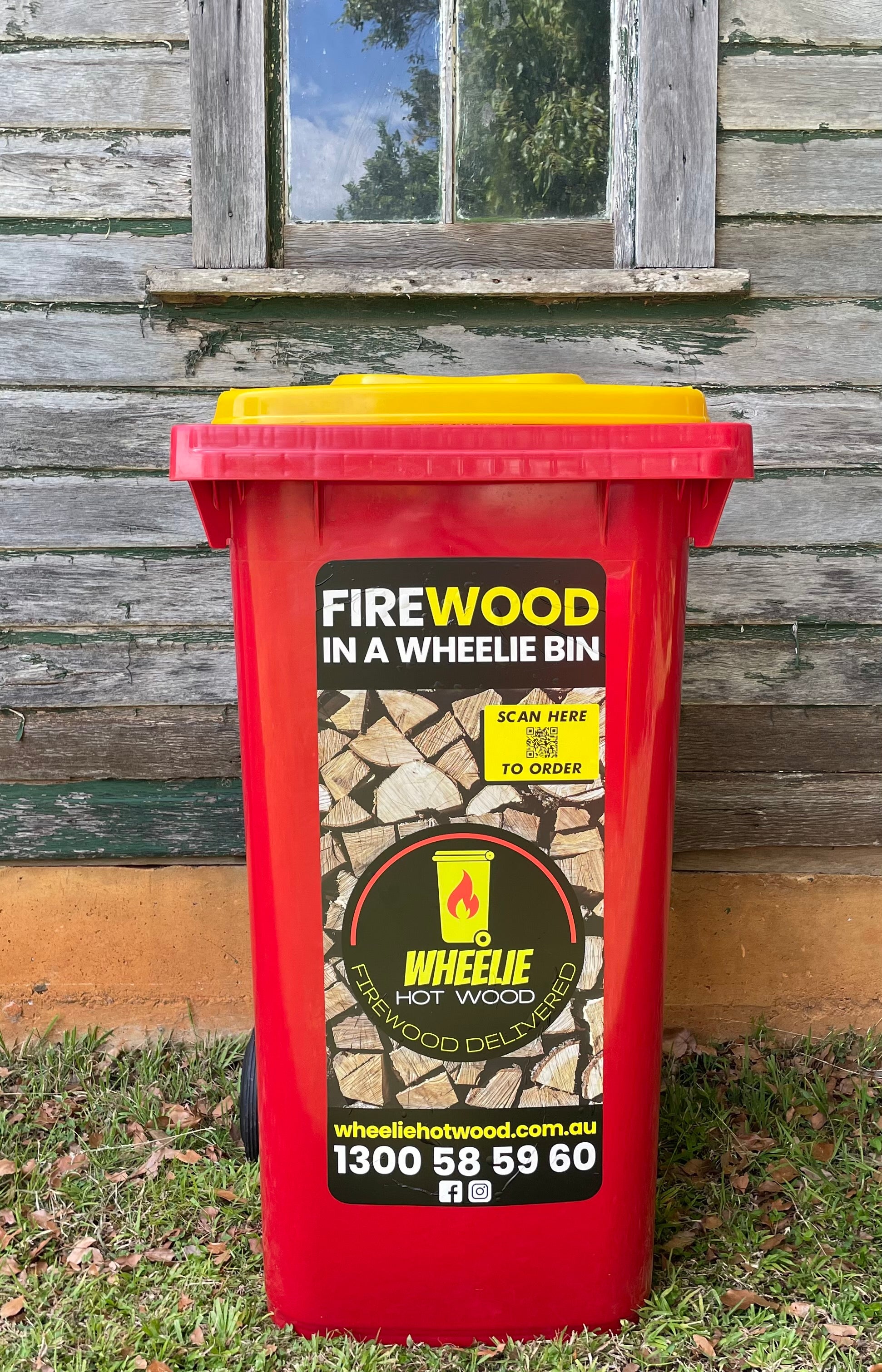 A red wheelie bin with a yellow lid, labelled "firewood in a wheelie bin", placed outside a house. Wheelie Hot Wood bins are available in Northern Rivers NSW and Gold Coast QLD