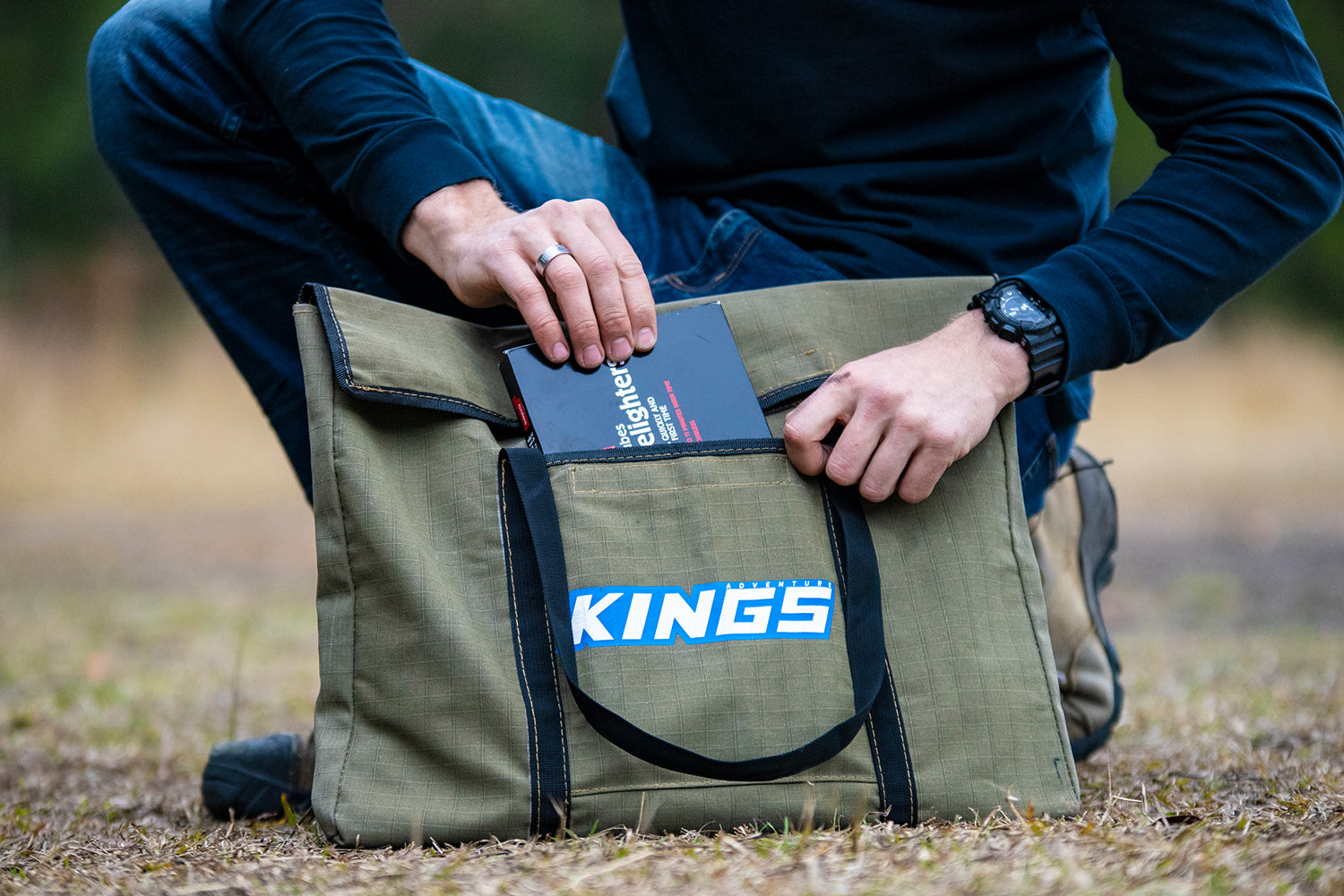KINGS Portable Firepit Canvas Bag, resting on the ground  with a man putting a box of firelighters into the front pocket