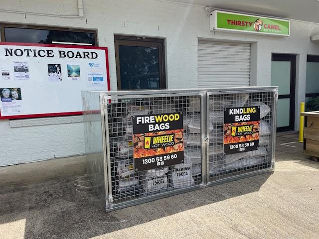 Wheelie Hot Wood Firewood  bag retail display cages sitting in front of a building with a notice board and Thirsty Camel bottle shop in Byron Bay. Wheelie Hot Wood delivers firewood in a wheelie bin in Northern Rivers NSW and Gold Coast, QLD.