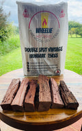 A stack of double split vintage ironbark wood in front of a bag labelled 'WHEELIE HOT WOOD - Double Split Vintage Ironbark 15kg' on a wooden surface with a green field in the background. Available in Northern Rivers NSW and Gold Coast QLD