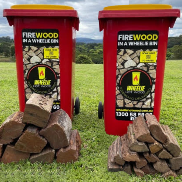 Two red wheelie bins labelled 'Firewood in a Wheelie Bin' with stacked firewood on a grassy field. One bin has a small pile of Standard Split firewood in front of it, and the other bin has a small pile of Double Split Firewood in front of it.