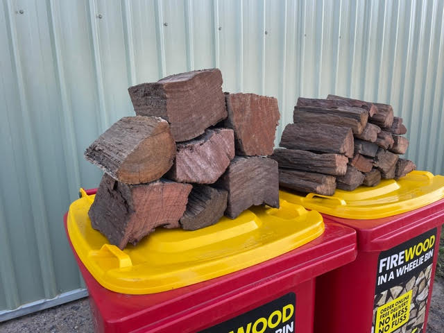 two red Wheelie Hot Wood bins with yellow lids sitting in front of a garage shed. one bin has a small pile of standard split firwood on it's lid, the other bin has a small pile of double split firewood on it's lid. The label on the bins says Firewood in a wheelie bin. Available in Northern Rivers NSW and Gold Coast, QLD.