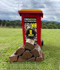 pile of standard split firewood in front of red Wheelie Hot Wood bin with yellow lid on a grassy hill. the label on the bin says Firewood in a wheelie bin. Available in Northern Rivers NSW and Gold Coast, QLD.