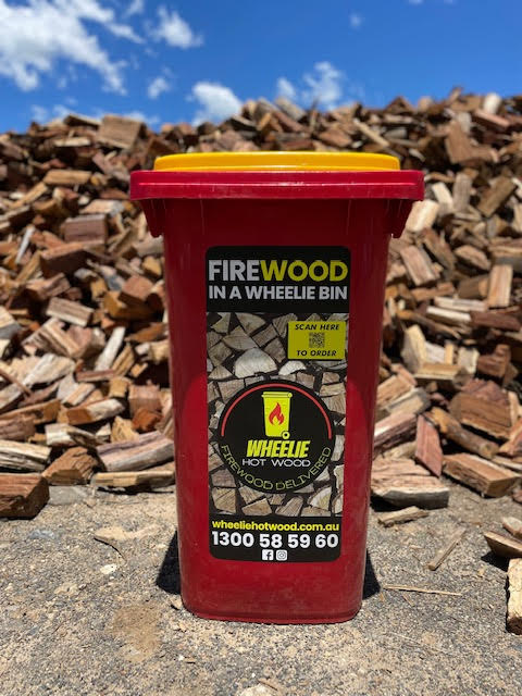 red Wheelie Hot Wood bin with yellow lid in front of a large pile of split firewood. the label on the bin says Firewood in a wheelie bin. Available in Northern Rivers NSW and Gold Coast, QLD.
