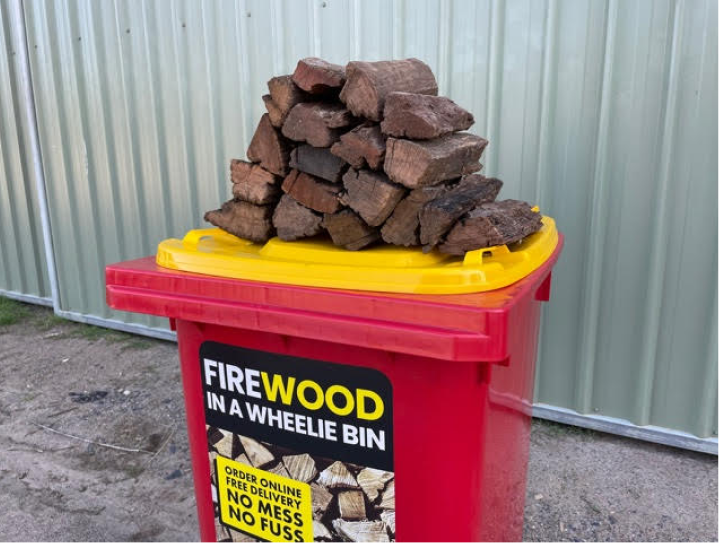 pile of double split firewood on top of the yellow lid of a red Wheelie Hot Wood bin sitting in front of a garage shed. the label on the bin says Firewood in a wheelie bin. Available in Northern Rivers NSW and Gold Coast, QLD.