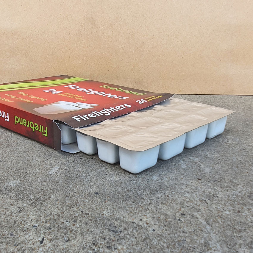 A box of Firebrand Firelighters containing 24 individual pop-out cubes, with red and green product packaging, lying flat on the ground with the tray pulled slightly out of the box