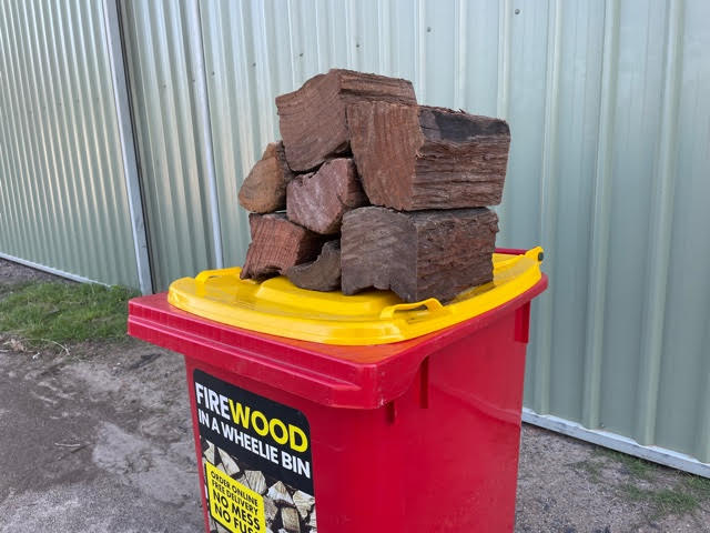 pile of standard split firewood on top of the yellow lid of a red Wheelie Hot Wood bin sitting in front of a garage shed. the label on the bin says Firewood in a wheelie bin. Available in Northern Rivers NSW and Gold Coast, QLD.
